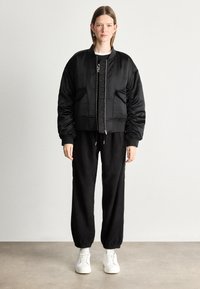 Czarna kurtka bomberka z wzmocnionymi ramionami i zapięciem na zamek, w połączeniu z luźnymi czarnymi spodniami i białymi sneakersami, stojąca na neutralnym tle.