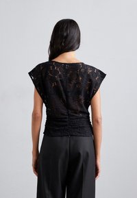 Haut en dentelle noire avec des manches courtes, présentant un motif floral et un design froncé à la taille. Porté avec un pantalon noir amplement ajusté.