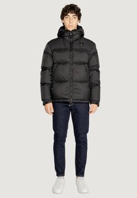 Giubbotto imbottito nero con cappuccio, dotato di chiusura a zip e due tasche laterali. Indossato sopra jeans blu scuro e scarpe da ginnastica bianche.