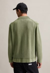 Knit green sweater with a collar, ribbed texture, and side stitching details, worn over black pants, viewed from the back.