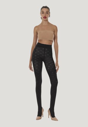 Kvinde stående iført en tan tube top, sorte mønstrede strømpebukser og nøgenfarvede høje hæle mod en ensartet lys baggrund.