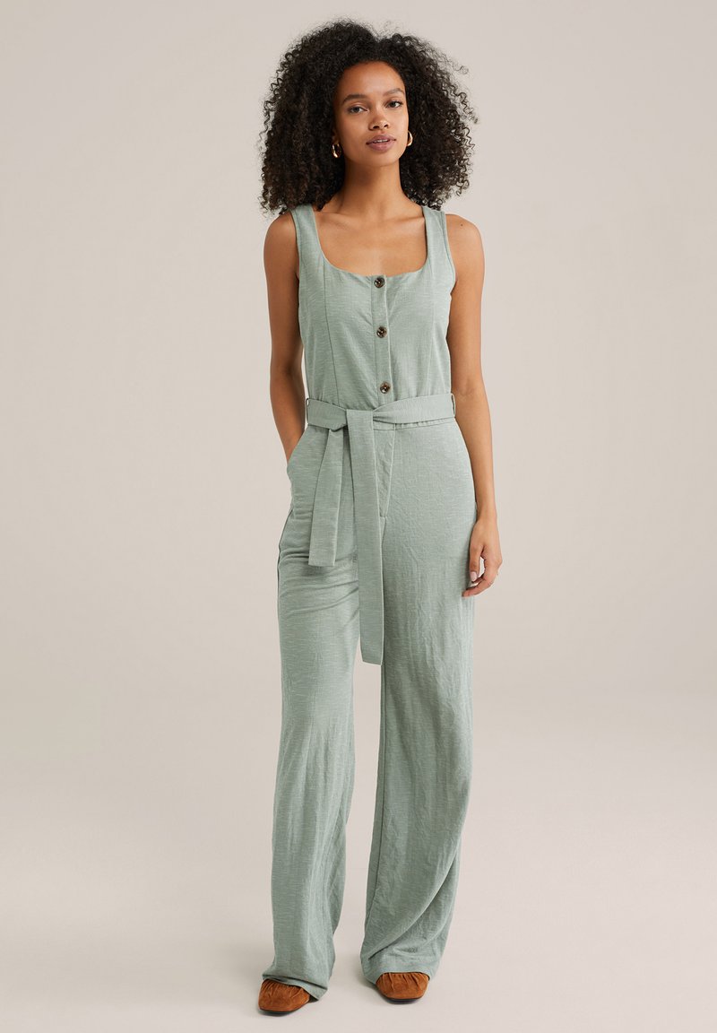 Ljusgrön linne jumpsuit med vida ben, fyrkantig halsringning och knappdetalj. Inkluderar ett tygbälte vid midjan.