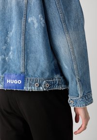 Šviesiai mėlyna džinsinė striukė su nusidėvėjusiu tekstūra, su mėlynu etiketė, kurioje užrašyta "HUGO", dviem sidabro spalvos snapeliais ir nusidėvėjusia apatine dalimi.