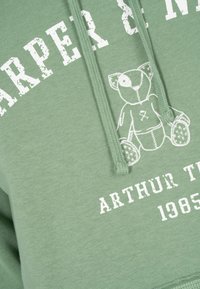 Sudadera verde con texto blanco "HARPER & M" y "ARTHUR T. 1985". Presenta un gráfico de osito de peluche y detalles de cordón. Textura de algodón suave.