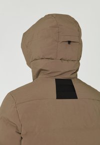 Brun huva puffer-jacka med en texturerad yta, med en svart panel på baksidan och en zip-ficka på sidan av huvan.