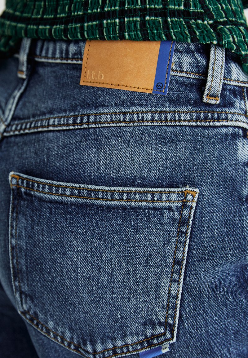 Blaue Jeans aus Denim mit einem cremefarbenen Lederpatch und einem blauen Akzentetikett auf der Rückseite. Verfügt über ein klassisches Fünf-Taschen-Design und orangefarbene Nähte.