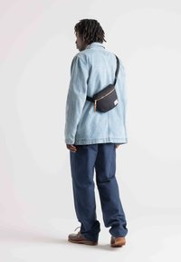 Denim jacket in light blue, navy pants, and brown shoes. Black waist bag with gold zipper detail worn crossbody. Simple, functional design.