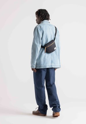 Leichte blaue Jeansjacke, marineblaue Hose und braune Schuhe. Schwarze Hüfttasche mit goldenen Reißverschlussdetails, diagonal getragen. Schlichtes, funktionales Design.