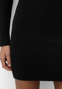 Černé žebrované šaty s dlouhými rukávy, které mají přiléhavý střih a jemnou texturu. Délka sukně sahá nad kolena a zdůrazňuje vertikální pruhy.