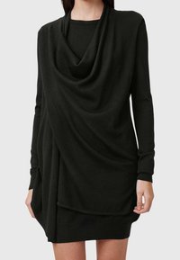 Femme portant une robe noire à manches longues avec un col bénitier drapé et une superposition asymétrique et ample.