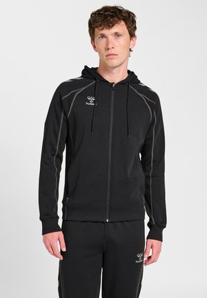 Jonge man draagt een zwarte hoodie met rits en bijpassende zwarte joggingbroek met witte stiksels en kleine logo's op borst en dij.