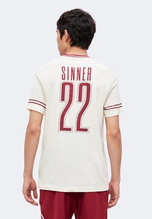 Mand iført en hvid sportstrøje med røde striber, "SINNER" og nummer 22 trykt på ryggen, kombineret med røde shorts.