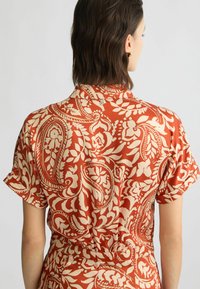 Roodbruin blouse met een crèmekleurig bloemen-paisleypatroon, korte mouwen en een kraag. Glad materiaal met een ontspannen pasvorm.