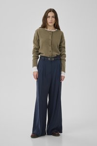 Frau, steht, trägt eine olivgrüne, geknöpfte Strickjacke, darunter ein weißes Hemd, weite marineblaue Hose und dunkle Schuhe.