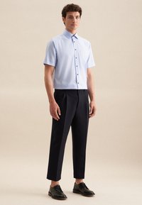 Camicia azzurra chiara a maniche corte con bottoni frontali e tasca, abbinata a pantaloni neri a pieghe e scarpe nere stringate su uno sfondo neutro.