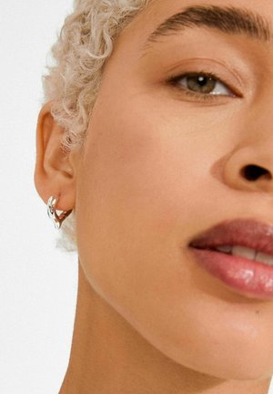 Hõbedast rõngakujuline kõrvarõngas, millel on keerduv disain. Suur plaan portreest, mis rõhutab sujuvat nahatooni ja loomulikku huulevärvi.