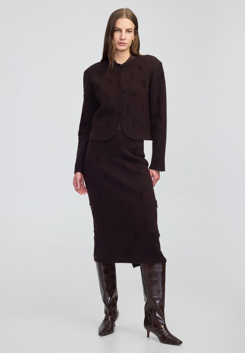 Bordeaux tweed outfit met een cropped jack met textuurstiksels en een bijpassende lange rok. Het model draagt glanzende bruine kniehoge laarzen.