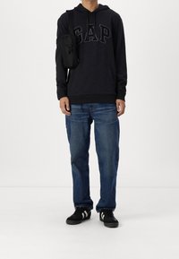 Sudadera con capucha negra con el logo "GAP", jeans de mezclilla azul y zapatillas negras con rayas blancas; pequeña bolsa cruzada negra.