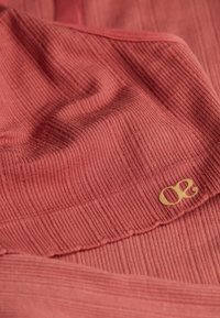 Tissu en terre cuite, doux et côtelé, avec un détail de logo doré. Le matériau est légèrement texturé, avec des lignes et des plis subtils.