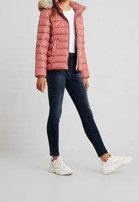 Pink puffer jacket with a fur-lined hood, featuring a zip front and flat quilting. Paired with dark skinny jeans and white sneakers.