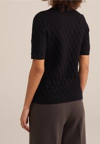 Person som bär en svart texturerad kortärmad tröja och grå byxor, visad bakifrån mot en enfärgad beige bakgrund.