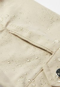 Teren de apă rezistent bej, cu o suprafață texturată, având picături de apă și logo-ul embosat "Joules". Include un detaliu cu capac de tip snap.