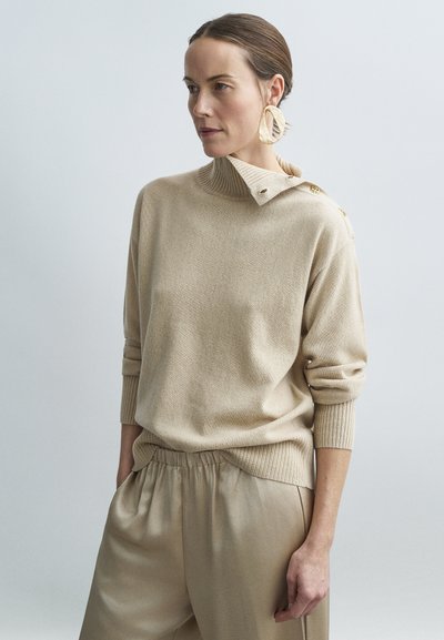 Donna con capelli tirati indietro che indossa un maglione beige a collo alto con bottoni, grandi orecchini astratti e pantaloni beige di satin larghi, con le mani in tasca.