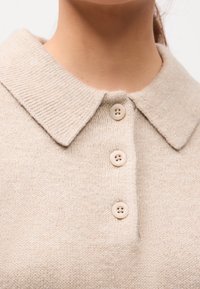Gros plan d'un pull beige en tricot avec un col pointu et trois boutons assortis sur le devant, porté par une personne.