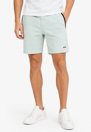 Homme portant un short gris clair avec une bande noire sur le côté et le logo "BOSS" sur la jambe gauche, des baskets blanches et un t-shirt blanc, debout avec une main dans la poche.