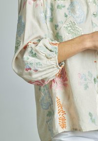 Blusa ricamata in tessuto beige con vivaci disegni floreali. Presenta maniche a palloncino con polsini arricciati e chiusura con bottoni.