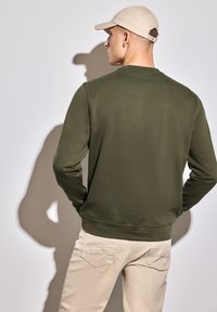 Olivově zelená mikina s dlouhými rukávy, ribbenovými manžetami a kulatým výstřihem, kterou doplňují světlé kalhoty. Hladká textura, jednoduchý design.