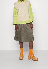 Frau in einem beigen ärmellosen Pullover über einem grünen Shirt, metallischem Faltenrock, orangen Socken und beigen Plateauschuhen, die vor einer weißen Wand steht.