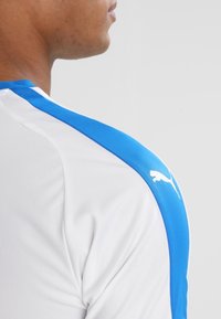 Man som bär en vit sporttröja med blå rand på axeln och vit Puma-logotyp, vänd sidledes mot en enkel bakgrund.