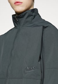 Nike jack in donkergrijs, met een hoge kraag, volledige rits en een geborduurd logo op de linkerborst. Gladde textuur, lichtgewicht materiaal.