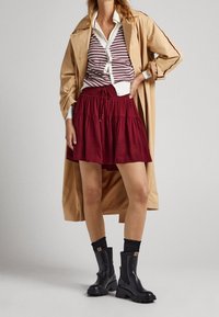 Kvinna klädd i beige trenchcoat, randig cardigan, röd kjol, svarta strumpor och kraftiga svarta stövlar, stående med handen på höften.