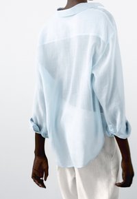 Camicia in lino leggera e trasparente di colore azzurro, con una vestibilità ampia, spalle scese e maniche arrotolate, caratterizzata da un colletto semplice e un orlo arrotondato.
