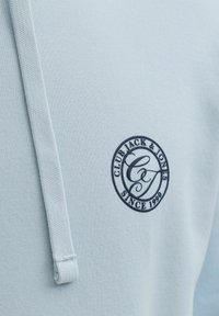 Vaaleansininen huppari, jossa on teksturoitu kangas, tummansininen pyöreä kirjailtu logo ja hupussa kiristysnauha.