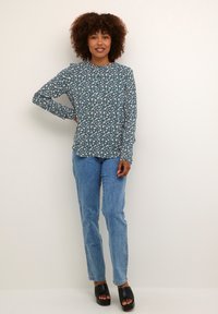 Blouse teal à manches longues avec motif floral blanc, associée à un jean droit bleu clair et des sandales à plateforme noires. Texture lisse.