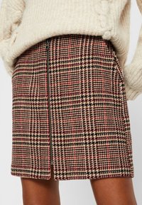 Vrouw gekleed in een wollen mini-jurk met een rood, zwart en beige houndstoothpatroon en een centrale zwarte rits, gecombineerd met een crèmekleurige gebreide trui.