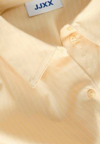 Chemise habillée pour hommes de couleur jaune clair avec de fines rayures blanches verticales et des boutons, présentant un col plié et une étiquette de marque JJXX à l'intérieur.