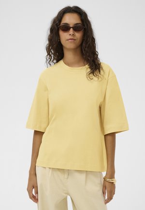 Woman with long curly hair wearing dark sunglasses, a loose yellow short-sleeve top, beige pants, and gold bracelets on her right wrist.