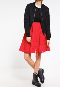 Černá bomber bunda, černé tričko, červená krajková sukně a černé šněrovací boty. Sukně má květinový vzor; bomber má žebrované manžety.