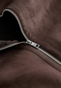 Interiorul unei jachete din suede maro, cu un fermoar argintiu. Textură moale, material neted și o căptușeală vizibilă din fleece în zona fermoarului.