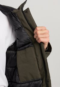 Giubbotto imbottito con zip frontale, caratterizzato da un esterno nero e lucido e una fodera interna verde oliva con tasca. Texture liscia.