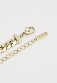 Χρυσή αλυσίδα βραχιόλι με πλεγμένο σχέδιο, κλιπ αστακού και ορθογώνια πινακίδα με το "LACOSTE" χάραξη. Λείος μεταλλικός φινίρισμα.