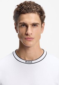 Νέος άνδρας με καστανά σγουρά μαλλιά που φοράει λευκό πουκάμισο με μαύρο ριγέ γιακά και κείμενο "BOSS" στον λαιμό, σε λευκό φόντο.