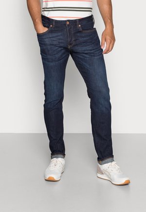 Homme portant un jean slim bleu foncé, des baskets blanches et grises, et un t-shirt rayé, debout les mains détendues.