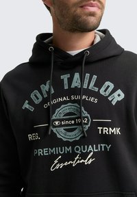 Schwarzer Kapuzenpullover mit einem Frontaufdruck, der die Texte "TOM TAILOR" und "PREMIUM QUALITY" zeigt, akzentuiert mit einem runden Logo. Zugbandverschluss am Hals.