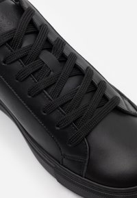 Baskets en cuir noir avec lacets plats tressés, texture lisse, bout arrondi et semelle en caoutchouc, avec un design minimaliste et des détails de couture.