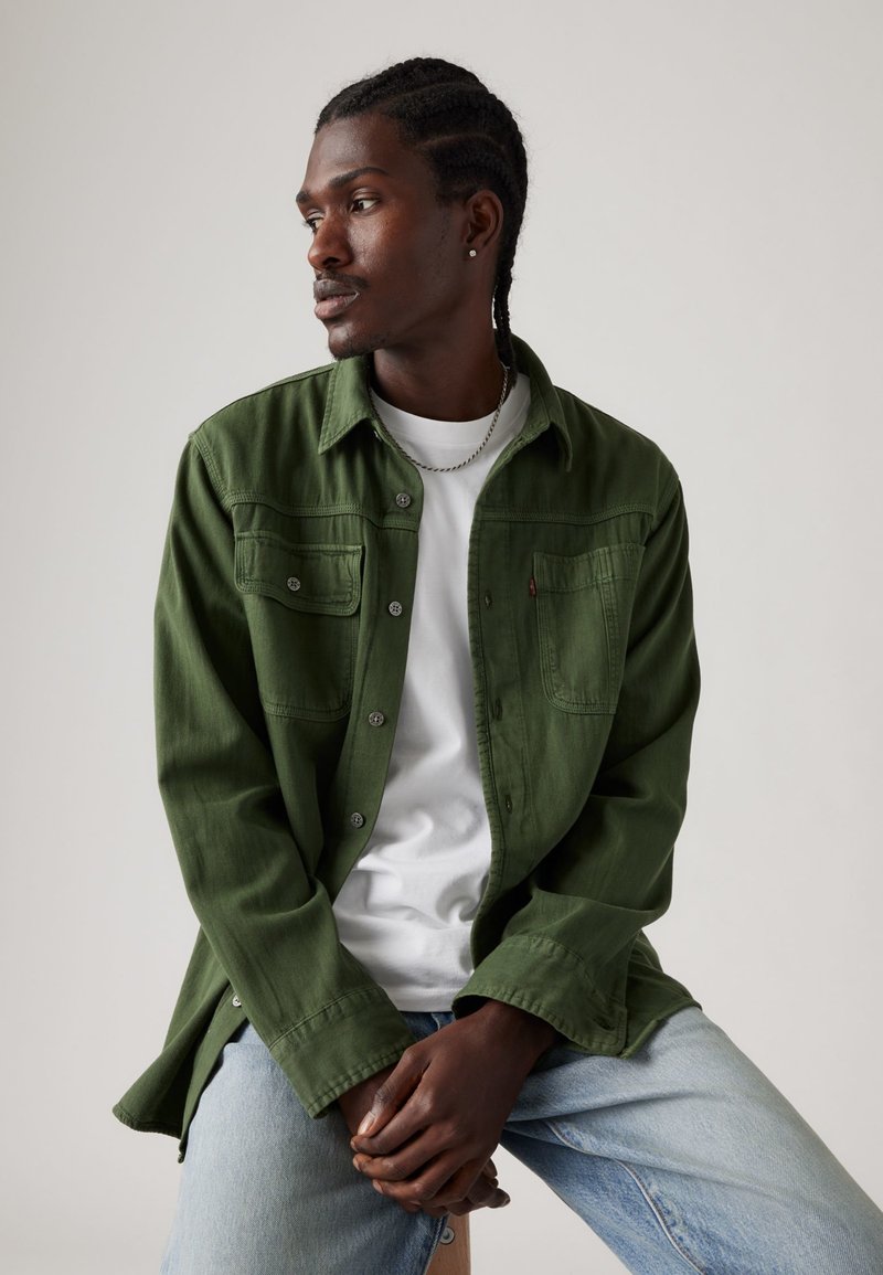 Chemise verte à boutons, avec deux poches poitrine, portée sur un t-shirt blanc. Le mannequin a les cheveux tressés et est assis. Les jeans sont bleu clair.